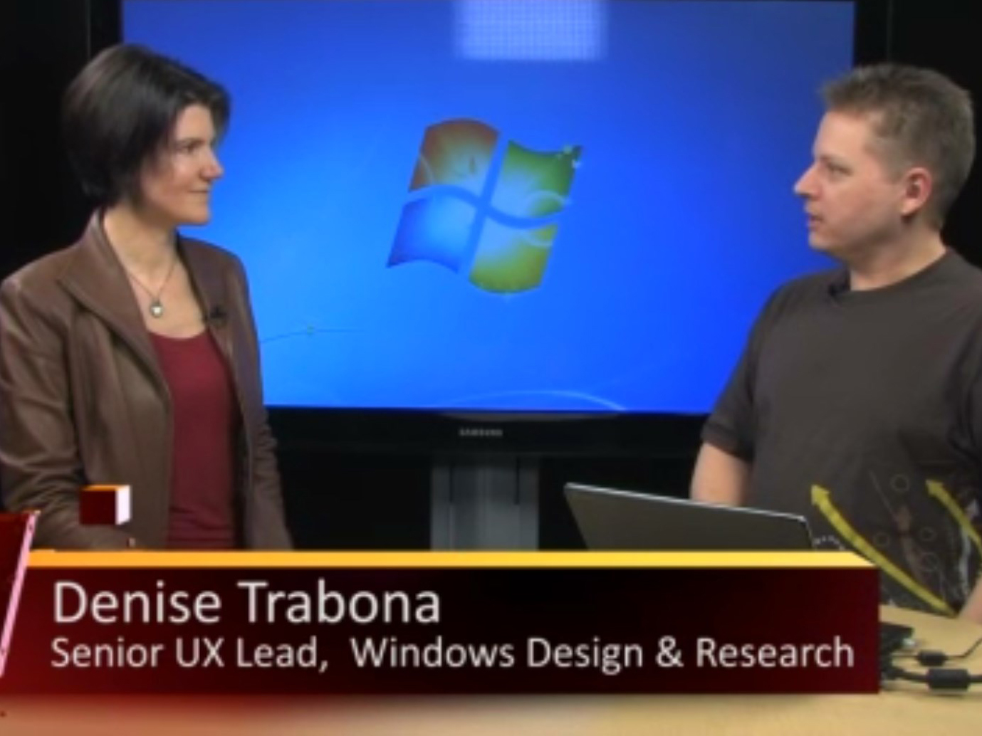 Denise Trabona and a presenter in front of a laptop with a big screen in the background featuring the Windows 7 default wallpaper